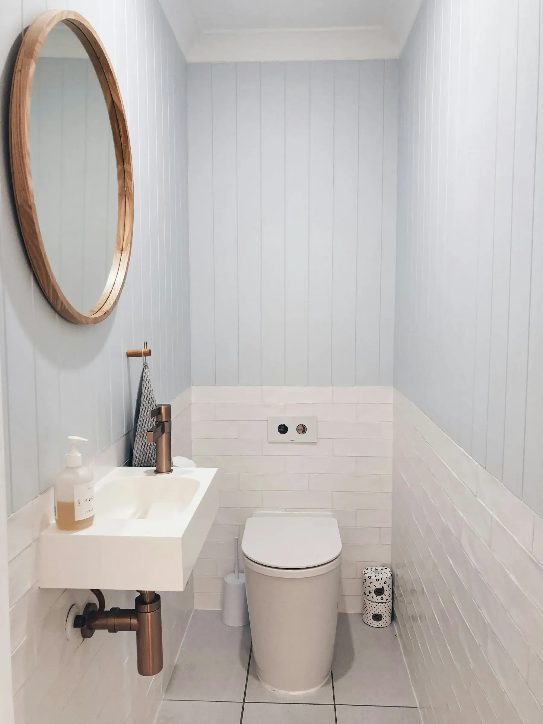 Modern bathroom interior showcasing impeccable white subway bathroom tiling in Wolverhampton, paired with light blue vertical wood panelling, a copper tap, and a round wooden mirror.