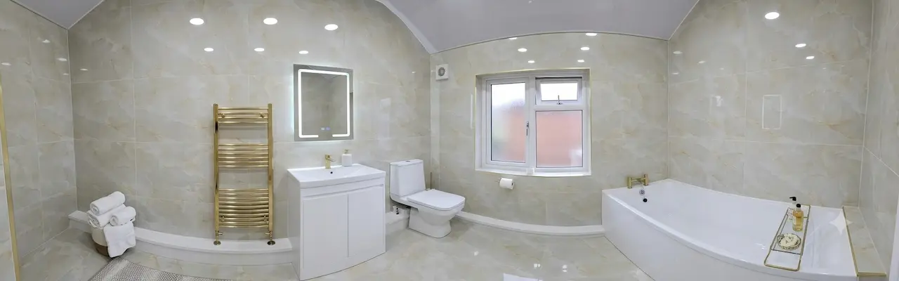 A panoramic, wide-angle photograph of a newly renovated, modern bathroom featuring polished beige marble-effect tiles on the walls and floor. The space includes a white vanity unit with a gold tap and illuminated mirror, a gold heated towel rail, a white toilet, a window, and a straight bathtub.